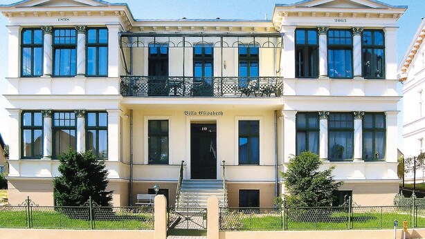 Un immeuble de deux étages, de style architectural classique, avec une entrée centrale dotée d’une porte noire et des balcons à chaque étage.