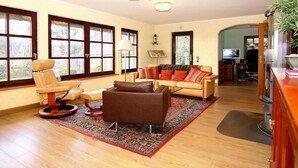 Amplia sala de estar con sofá, sillón y mesa de centro. Cuenta con ventanas amplias, televisor y una planta.