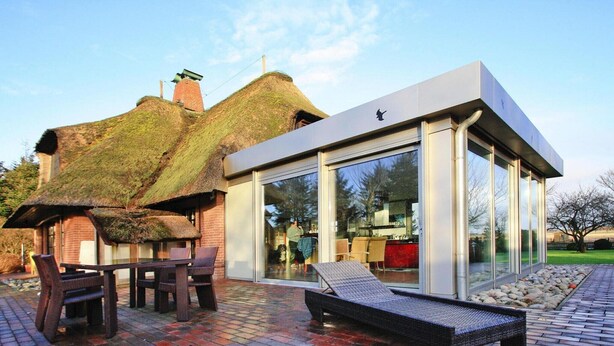 Una casa moderna con un cerramiento de vidrio, techo de paja y patio de ladrillo con mobiliario exterior.