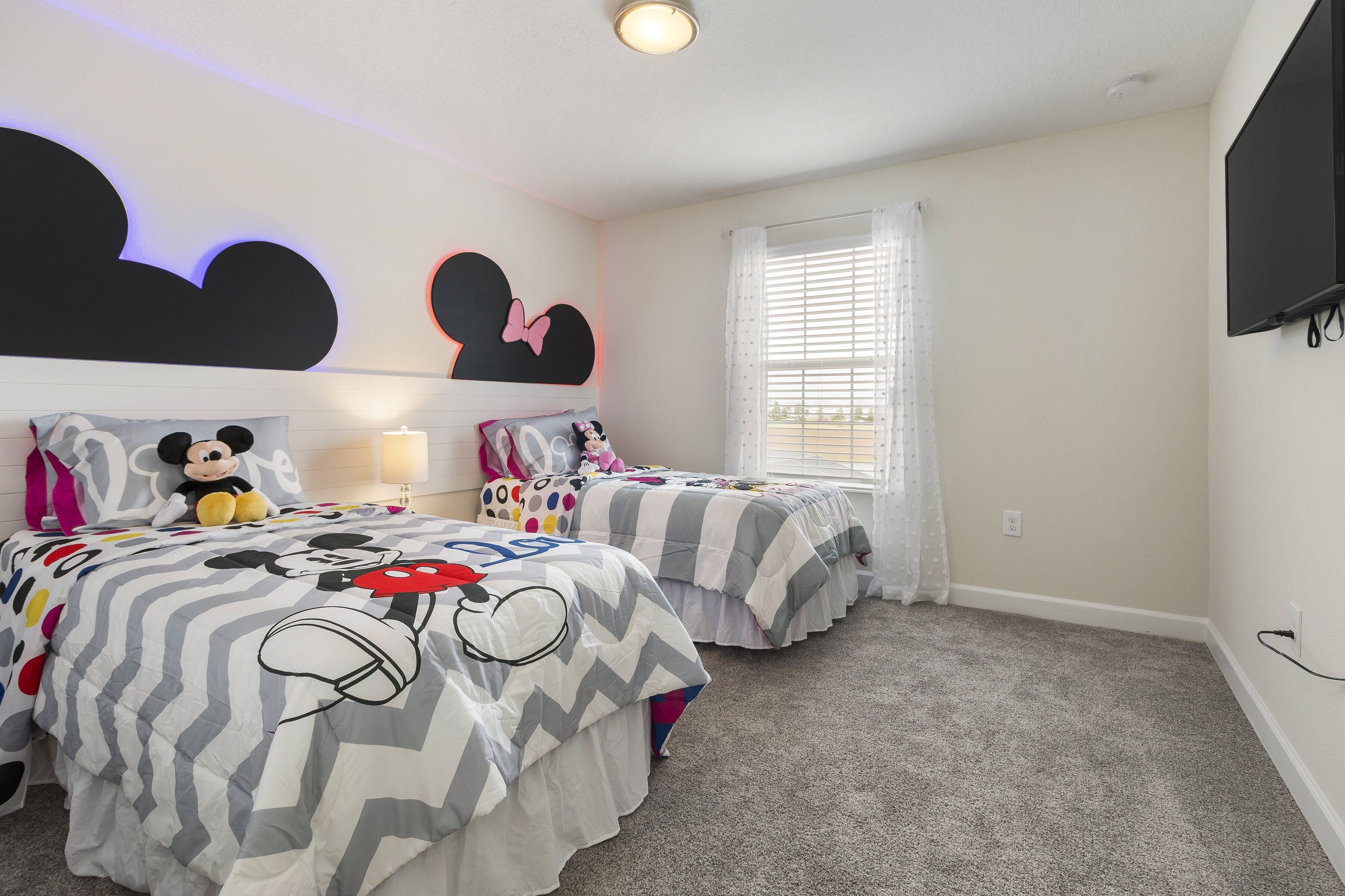 Ein Schlafzimmer mit zwei Betten, beide mit Bettwäsche im Mickey-Mouse-Design. An der Wand hängt eine große Mickey-Mouse-Dekoration, ein Fenster mit Jalousien und ein an der Wand befestigter Fernseher.