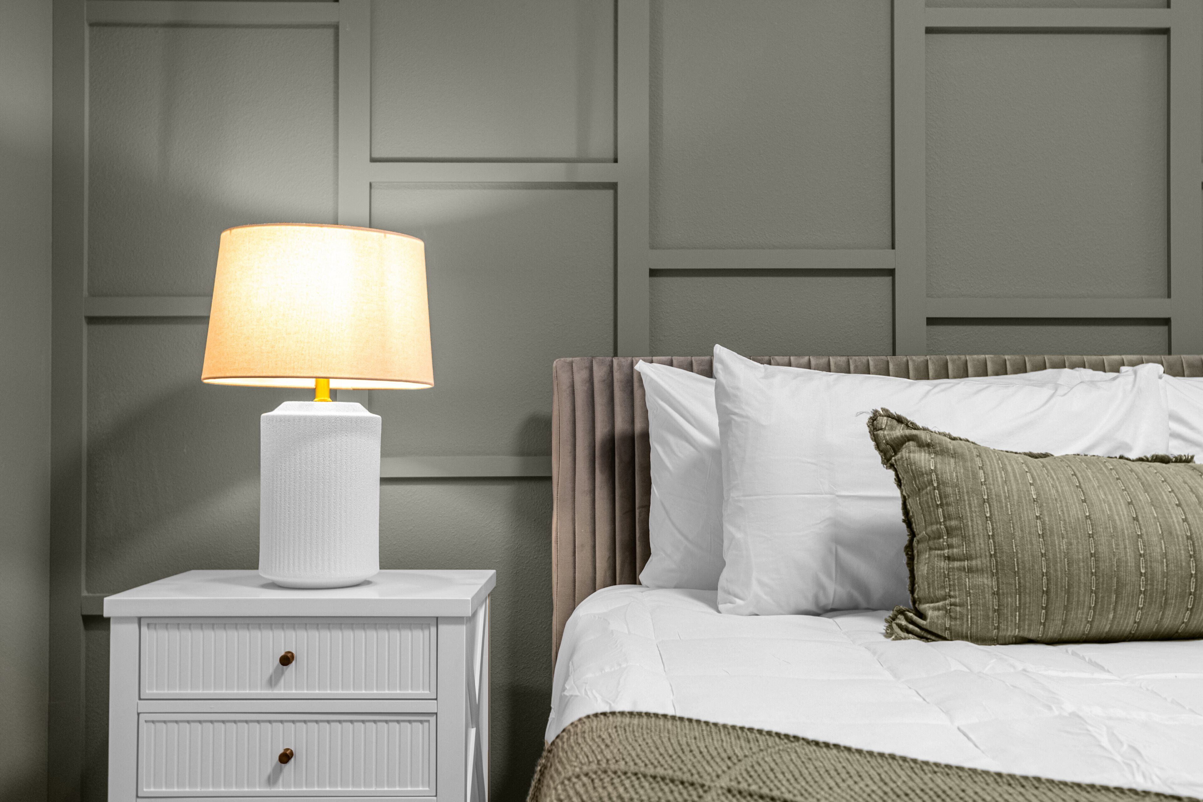A neatly made bed with white linens and a single green pillow, a white bedside table with a lamp, and a wall with paneling.