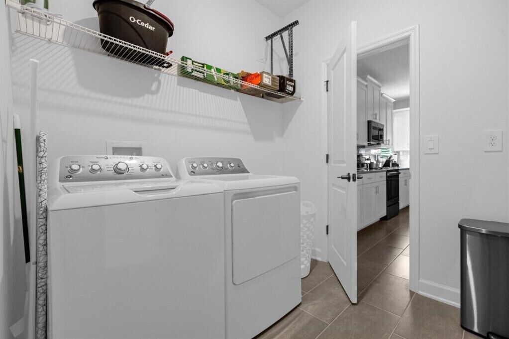 A laundry room with two white front-loading washers and dryers, a shelf with a potted plant, and a door leading to a kitchen area.