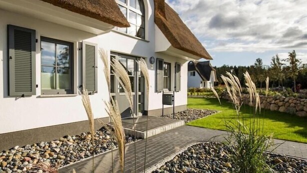 A modern house with a thatched roof, large windows, and a well-maintained garden with rocks and plants.