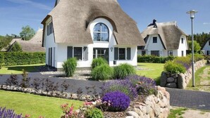 A white house with a thatched roof, surrounded by a well-maintained garden with various plants and a stone wall.