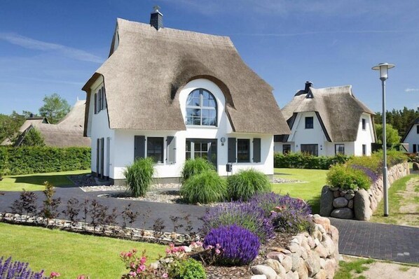 A white house with a thatched roof, surrounded by a well-maintained garden with various plants and a stone wall. - Luxushaus Boddenküste mit Lagune und Sauna (Fuhlendorf)