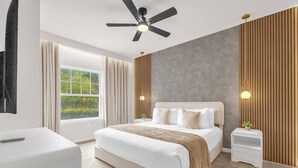 A bedroom with a bed, bedside tables, a ceiling fan, a window, and a view of greenery.