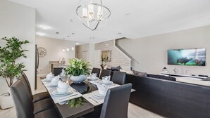 A modern dining area with a long table set for four, a sofa, a TV, and a staircase. - Last Minute Disney Getaway! Townhouse with Private Pool. Book Now! #65787 (Orlando)