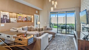 A spacious living room with a large sofa set, a coffee table, and a television. There is a sliding glass door leading to an outdoor patio area. - Last Minute Disney Getaway! Luxury Villa with Private Pool. Book Now! #66003 (Orlando)