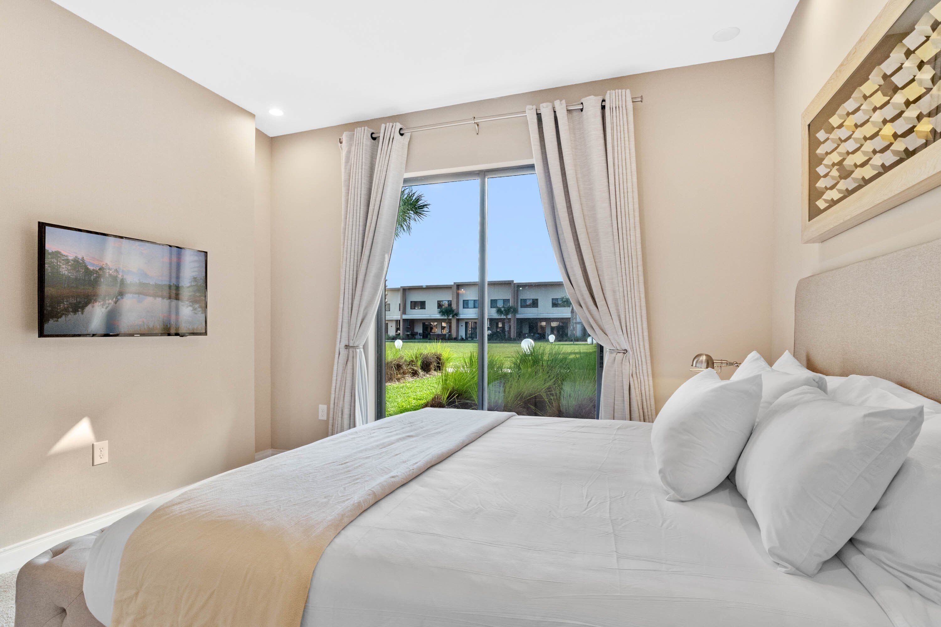Une chambre d’hôtel moderne dotée d’un grand lit, d’un téléviseur à écran plat fixé au mur et offrant une vue sur un jardin aménagé, visible à travers une grande fenêtre.