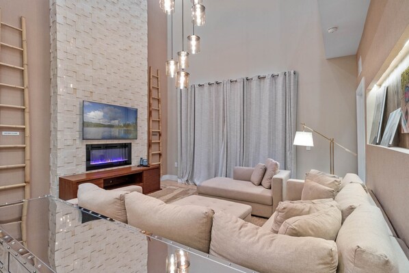 A modern living room with a flat-screen TV, beige sofas, a glass coffee table, and a fireplace. - Last Minute Disney Getaway! Townhouse with Private Pool. Book Now! #66049 (Orlando)