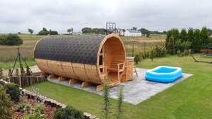 A wooden barrel-shaped structure with a ladder leading up to a platform, surrounded by a grassy yard and a blue inflatable pool. - Large, 2-story holiday home, sauna, jacuzzi (Mielenko)