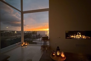 Ein Zimmer mit großem Fenster und Blick auf eine Stadtlandschaft bei Sonnenuntergang, einem Kamin und einem Tisch mit Kerzen.