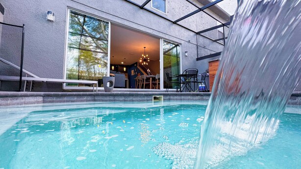 Une piscine agrémentée d’une cascade, un patio couvert avec des sièges d’extérieur, et un bâtiment doté de grandes fenêtres laissant entrevoir l’intérieur.