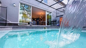 Une piscine agrémentée d’une cascade, un patio couvert avec des sièges d’extérieur, et un bâtiment doté de grandes fenêtres laissant entrevoir l’intérieur.