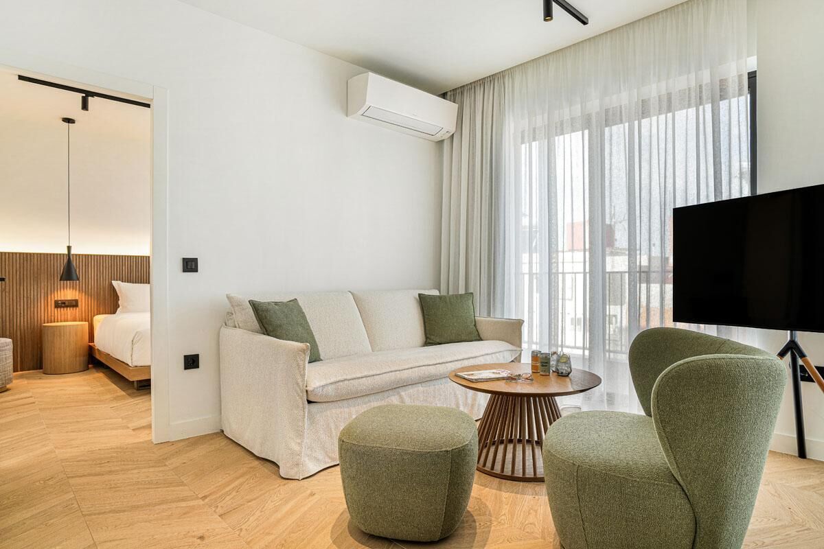 Een moderne woonkamer met een bank, poef en fauteuils, een flatscreen televisie en een groot raam met dunne gordijnen.
