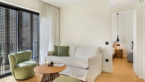 Una sala moderna con sofá, sillón y mesa de centro, ventanas grandes con persianas y suelo de madera.