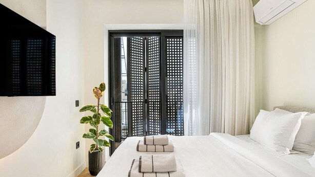 Una habitación de hotel con una cama, una planta en maceta, un televisor montado en la pared y una ventana con cortinas.