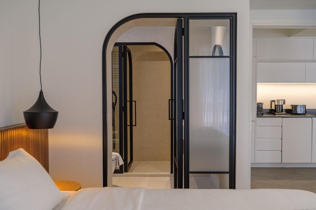 Moderni hotellihuone, jossa on sänky, yöpöytä, lamppu, lasiseinäinen kylpyhuone ja pieni keittiönurkkaus.