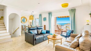 Ein großzügiges Wohnzimmer mit einer großen Couch, einer kleineren Couch und einem runden Couchtisch. Eine Treppe führt zu einer oberen Etage, und ein Balkon bietet Blick auf das Meer.