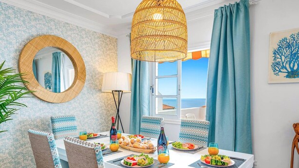 Ein Essbereich mit einem weißen Tisch, der für das Frühstück gedeckt ist, einer Weiden-Pendelleuchte und Meerblick durch ein Fenster.