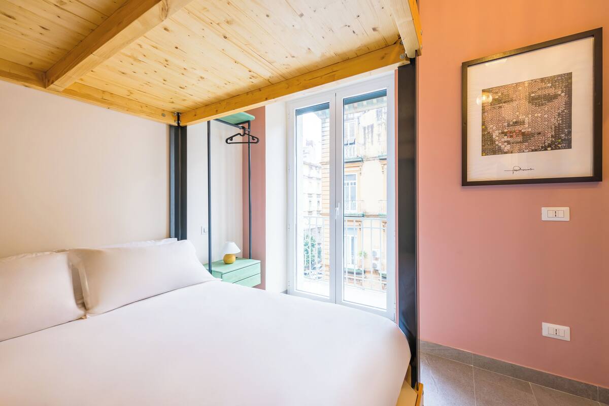 A bedroom with a bed, a window, a framed artwork, and a green nightstand.