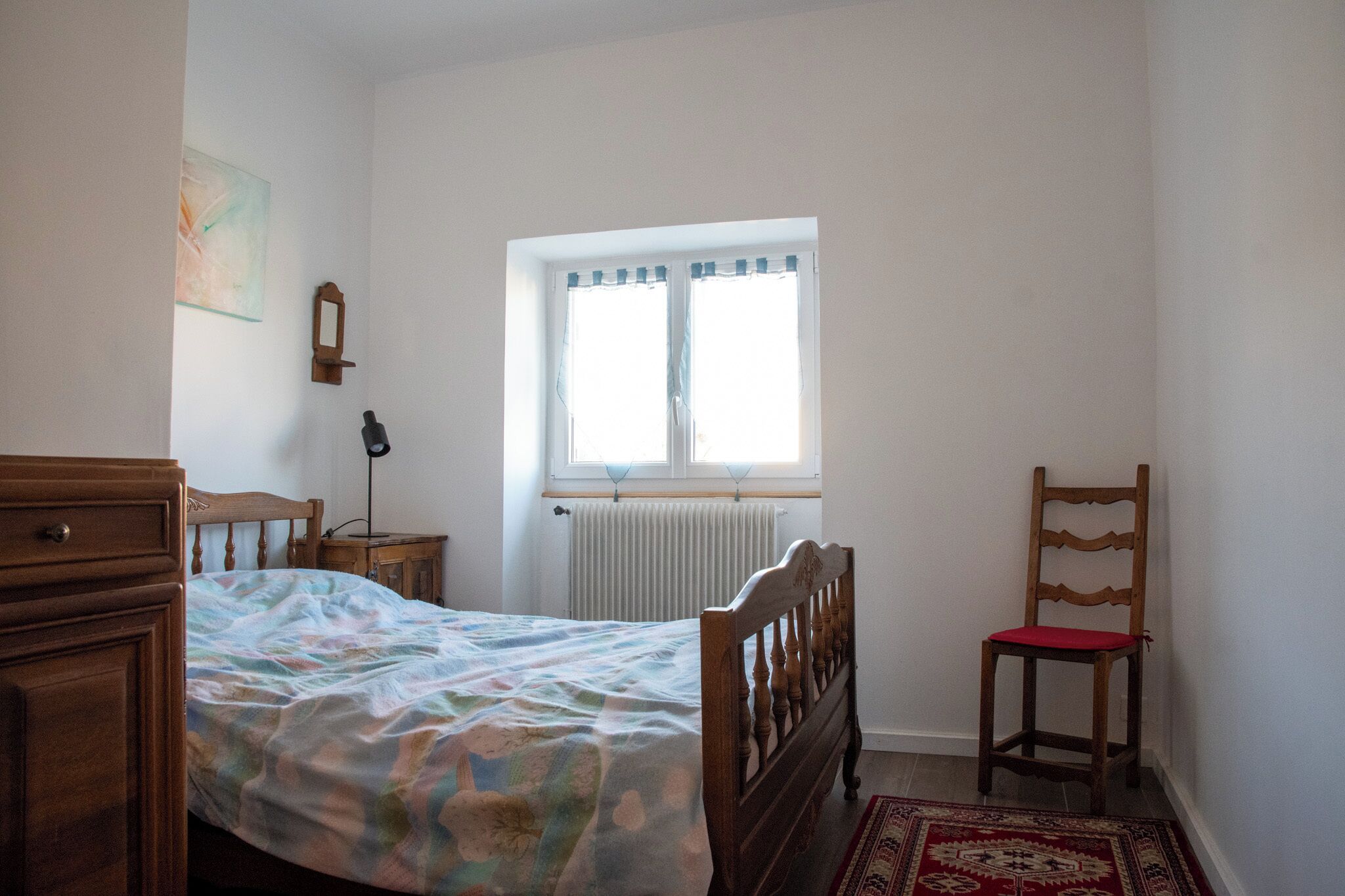 Ein Schlafzimmer mit Bett, einem Holzstuhl, einem Nachttisch, einem Fenster mit Vorhängen, einem Heizkörper und einem an der Wand befestigten Spiegel.