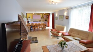 Un salon avec un téléviseur, un canapé et une table basse. Au fond, on aperçoit une cuisine avec des placards et un coin repas.