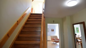 Un escalier en bois avec une rampe, menant à une pièce où l’on trouve une croix au mur, une chaise et une table.