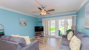A living room with two grey sofas, a glass coffee table, a ceiling fan, a TV, and a sliding glass door leading to a patio.