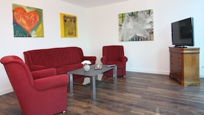 A living room with a red sofa, a red armchair, a coffee table, and a television on a wooden cabinet. There are three paintings on the wall.