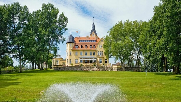 A large, historic building with a prominent tower, surrounded by lush green lawns and a fountain in the foreground.