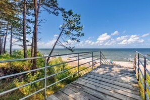A wooden boardwalk leading to a beach with a railing, surrounded by trees and a clear sky.