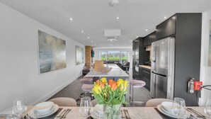 Een moderne keuken met een centraal eiland, donkere kastjes en roestvrijstalen apparatuur. Er is een eethoek met een tafel voor zes personen, en een groot raam met uitzicht op de omgeving.
