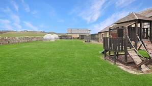 A grassy outdoor area with a wooden play structure, a stone wall, and a building with a sloped roof in the background.