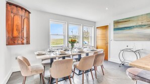 A dining area with a table set for six, large windows, a wooden cabinet, and a painting on the wall.