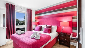 Una habitación de hotel moderna con una cama grande, paredes a rayas rojas y blancas, una ventana con cortinas y una mesita de noche con una lámpara.