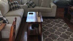 A living room with a sofa, a coffee table, a lamp, and a ceiling fan. There are two windows and an air conditioning unit. - Byron's House At Secret Beach. (San Pedro)