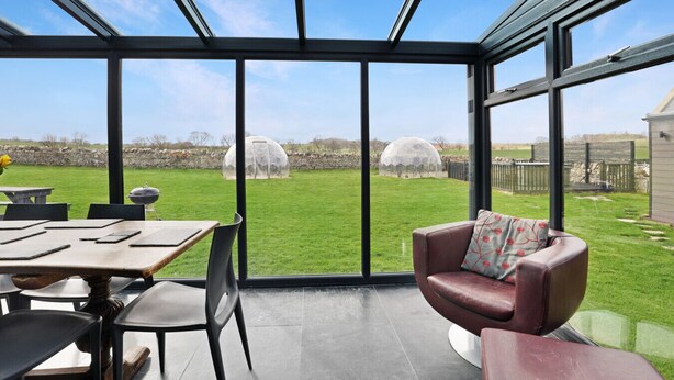 Une terrasse moderne dotée d’une véranda en verre, d’un espace repas et d’un coin salon confortable offrant une vue sur un jardin et une serre.
