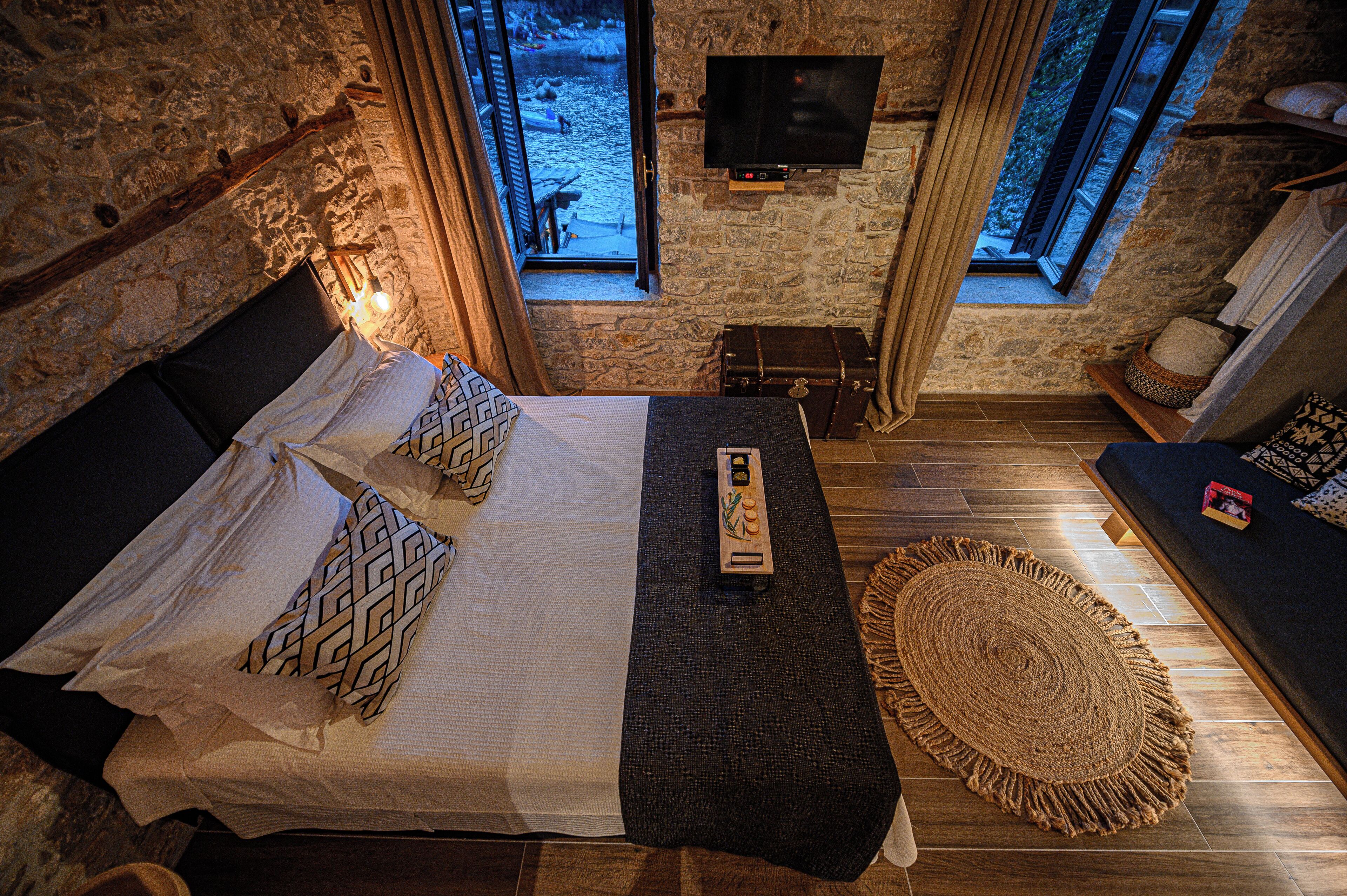 A bedroom with a stone wall, a bed with patterned pillows, a remote control, a woven rug, and a television on the wall.