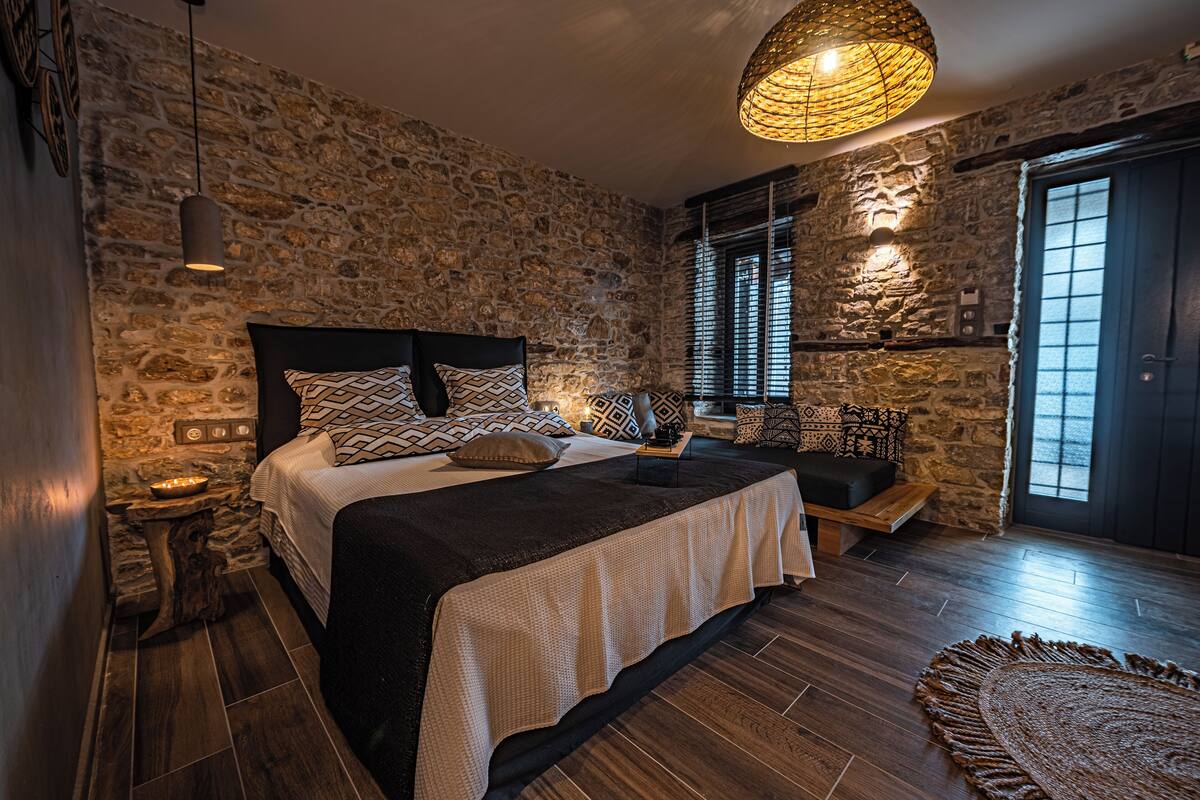 Une chambre avec un mur en pierre, un lit agrémenté d’oreillers à motifs, un luminaire suspendu et un banc en bois.