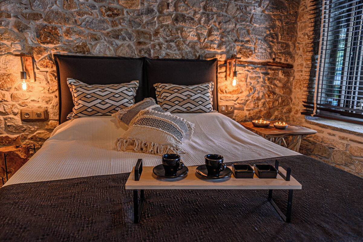 Une pièce chaleureuse avec un mur en pierre, un lit agrémenté d’oreillers à motifs, une table de chevet avec une lampe et un plateau contenant des tasses et des bols.