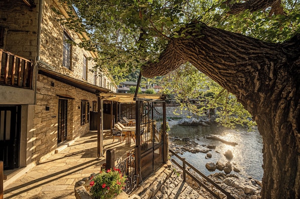 A stone building with a balcony overlooking a river, featuring outdoor seating and a view of the water.