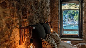 A cozy room with a stone wall, a window with a view of boats and greenery, and a bed with patterned pillows. - Cleopatra's Miramare (Zagora-Mouresi)