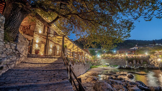 A stone staircase leading to a building with a balcony, a river in the background, and a bridge over the river. - Cleopatra's Miramare (Zagora-Mouresi)