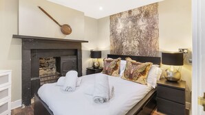 A bedroom with a fireplace, a bed with white linens, two bedside lamps, and a large abstract painting on the wall.