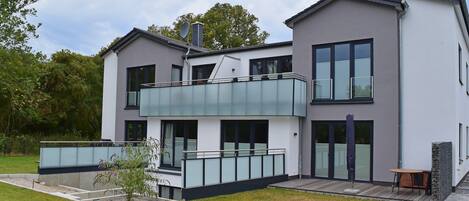 Une maison moderne à deux étages, dotée d’un balcon, de grandes fenêtres et d’un petit espace extérieur aménagé pour s’asseoir.