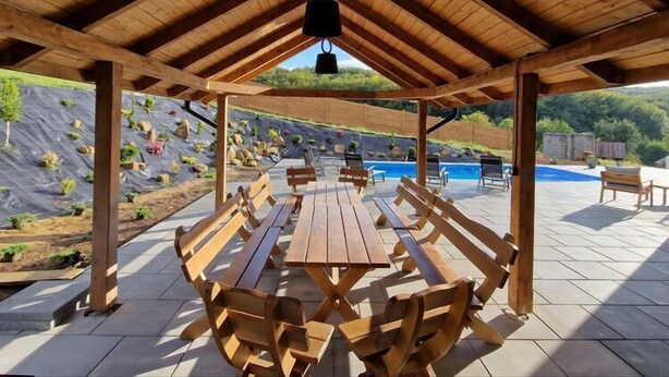 Een terras buiten met houten tafels en banken, een zwembad en uitzicht op een aangelegde tuin.