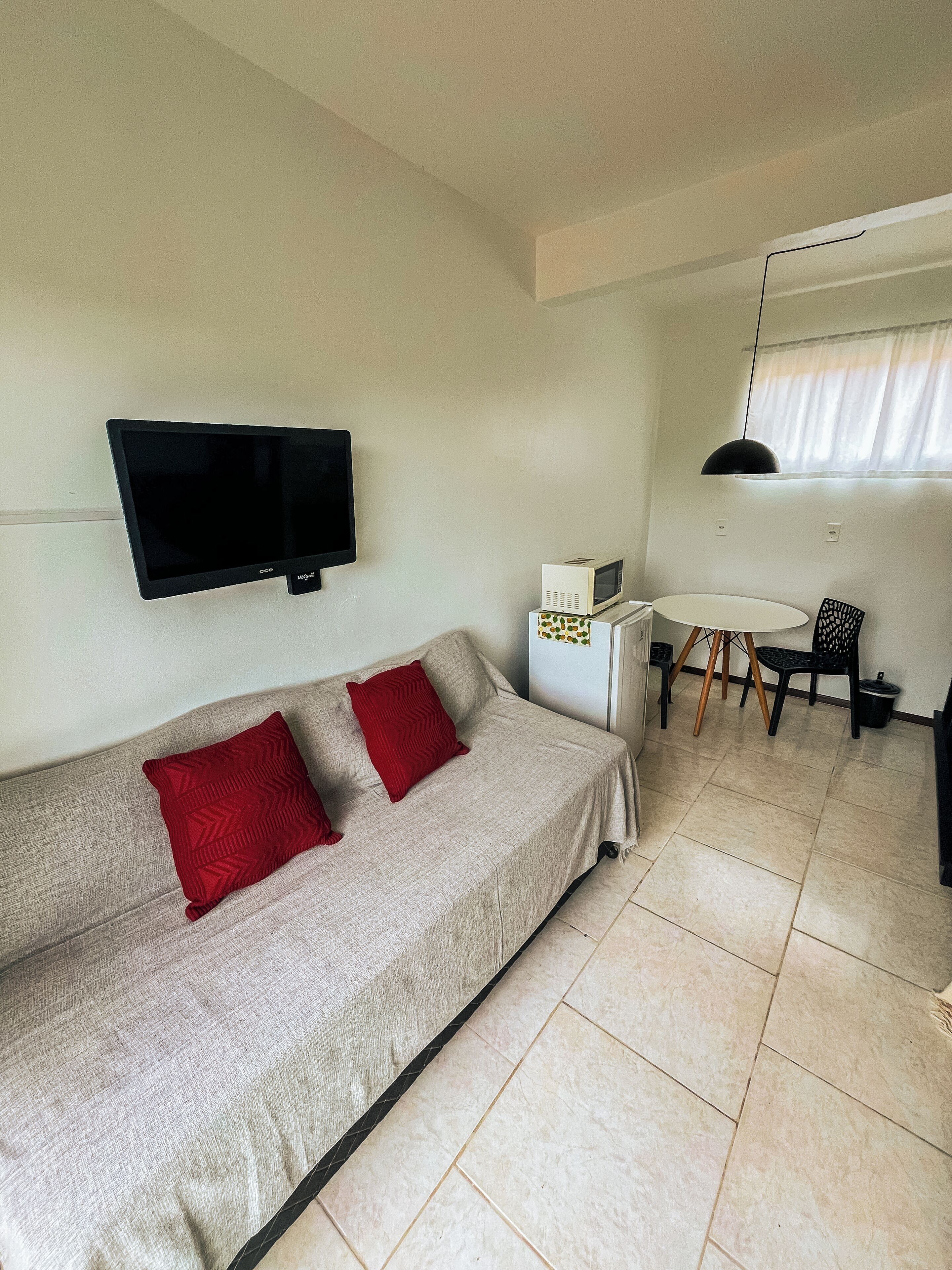 A living area with a sofa, two red pillows, a flat-screen TV, a microwave, a small table with chairs, and a pendant light.