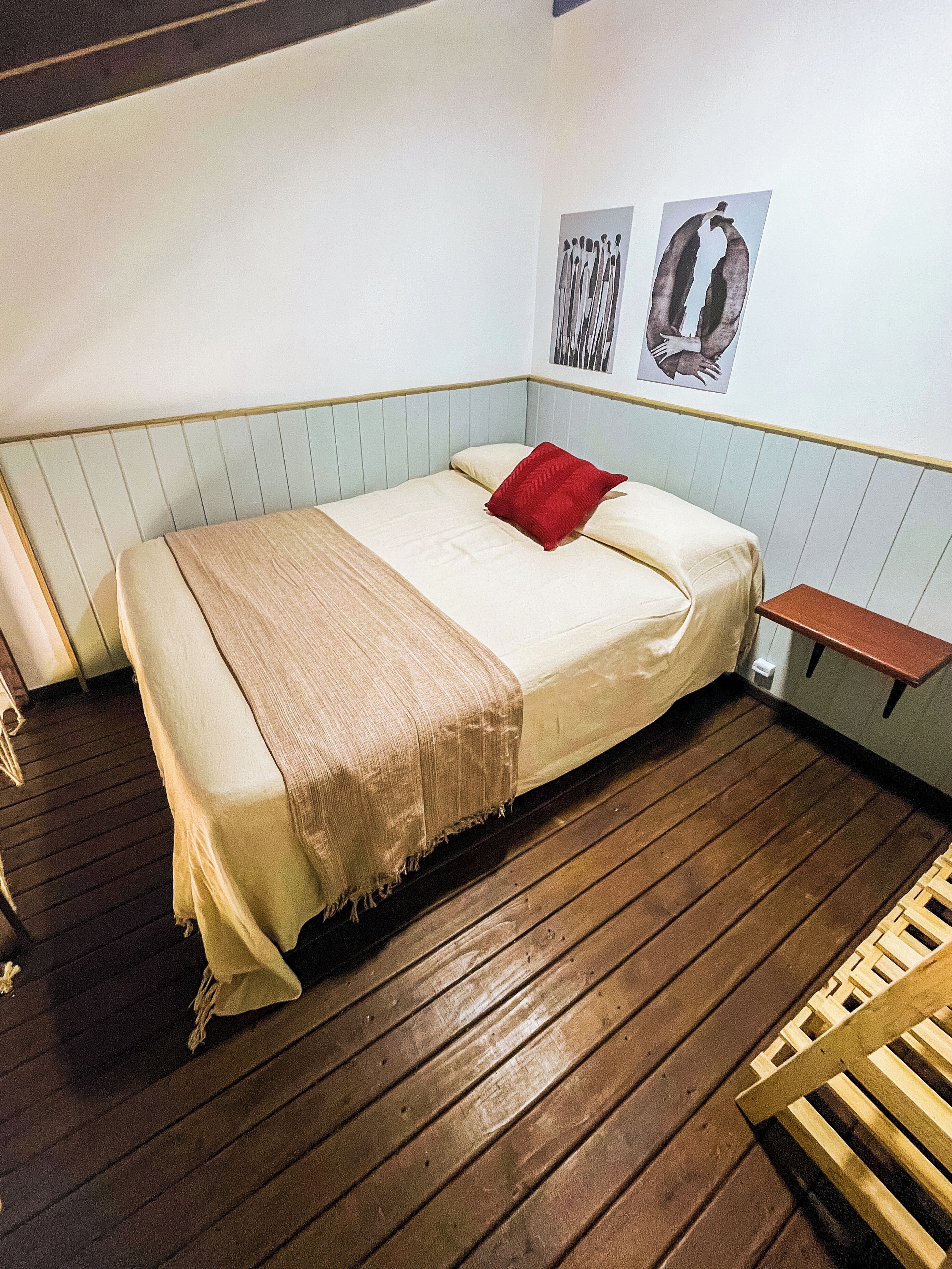 A single-bed room with a wooden floor, a small bedside table, a red pillow, and a wall-mounted picture.