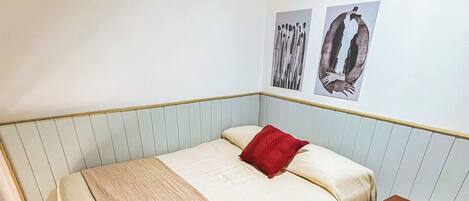 A single-bed room with a wooden floor, a small bedside table, a red pillow, and a wall-mounted picture.